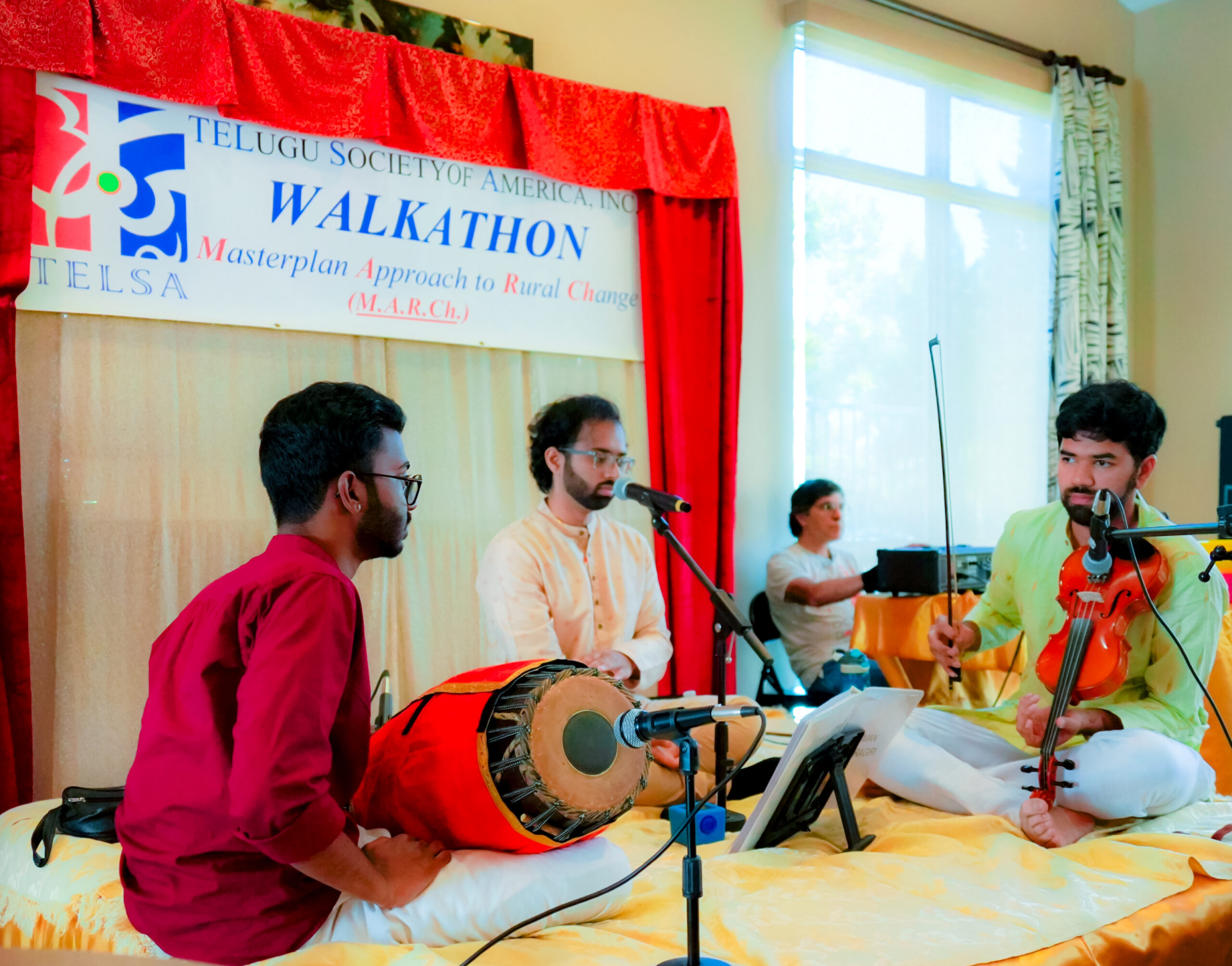 Three musicians performing traditional Indian classical music on a stage. One man plays the mridangam, another sings into a microphone, and a third plays the violin. A banner for the Telugu Society of America (TELSA) Walkathon is visible in the background behind red curtains.