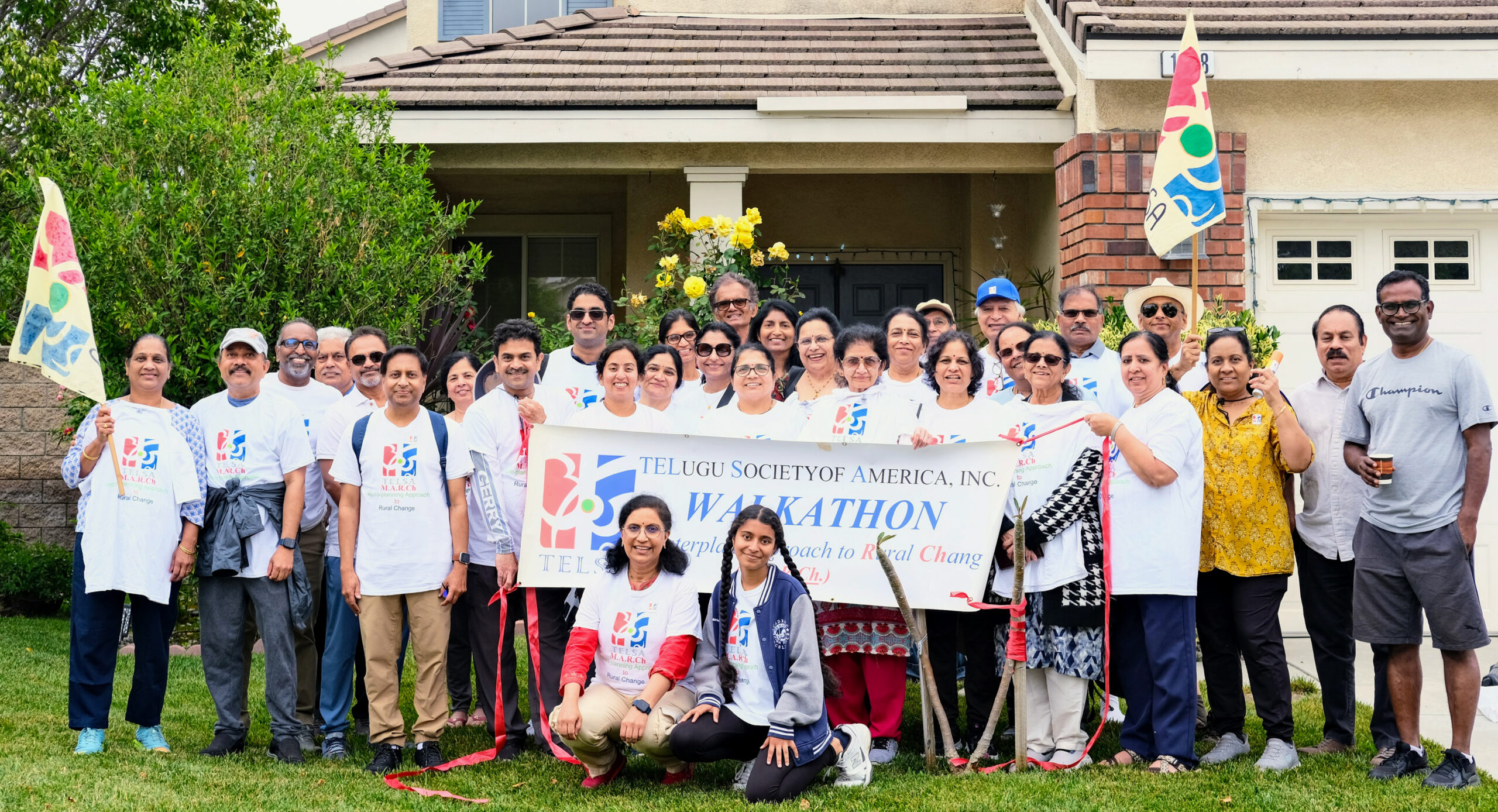 A large group of people of various ages wearing white TELSA branded t-shirts standing together in front of a suburban house. They are holding a large banner that reads "TELUGU SOCIETY OF AMERICA, INC. WALKATHON" and some are holding yellow and white flags.